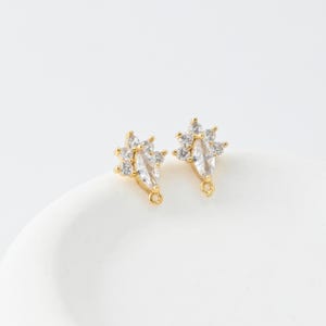 May include: A pair of gold-colored stud earrings. Each earring features a central marquise-cut clear crystal, surrounded by smaller, round clear crystals. The earrings have a post and loop for attachment.