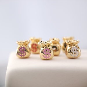 Puede incluir: Seis abalorios de bolsa de dinero de color dorado con detalles en esmalte rosa y blanco. Los abalorios presentan un signo de dólar en la parte delantera.