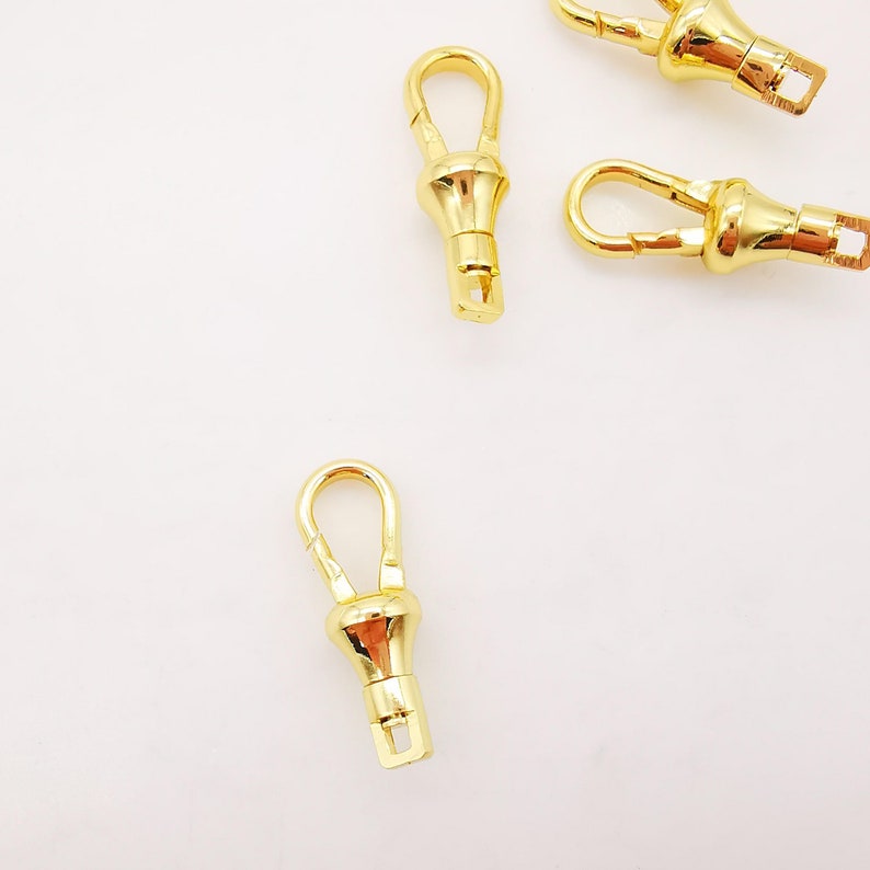 Puede incluir: Cuatro cierres giratorios de metal dorado con un bucle en un extremo y un gancho en el otro. Los cierres est&aacute;n dise&ntilde;ados para sujetarse a bolsos, carteras u otros art&iacute;culos.