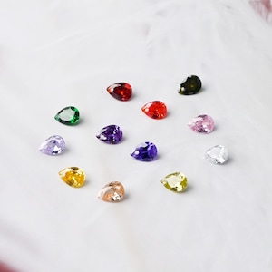 May include: A collection of teardrop-shaped gemstones in various colors, including red, green, purple, and yellow. The gems are arranged on a white surface, showcasing their facets and brilliance. Ideal for jewelry making or crafting.