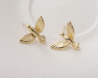 May include: A pair of gold-colored stud earrings in the shape of flying birds. The earrings have detailed wing and tail feather designs. The birds are facing opposite directions, creating a symmetrical look. The earrings are on a white surface.