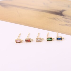 May include: Five gold earring posts with different coloured rectangular gemstones. The gemstones are pink, red, clear, green, and blue.