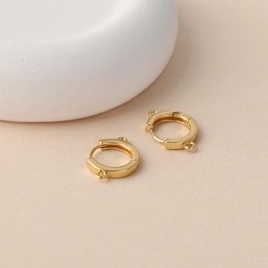 May include: A pair of small, polished gold-tone hoop earrings, each featuring a smooth circular band and a tiny integrated loop at the bottom for attaching charms or pendants. These circular jewelry components have a hinged closure mechanism.