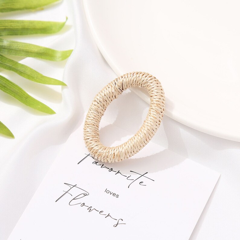 May include: A woven, oval-shaped napkin ring in a natural beige color. The ring is made of tightly woven fibers, creating a textured surface. The background includes a white plate, a white cloth, and a card with the words "favorite loves flowers".