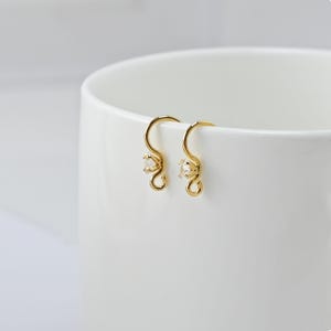 May include: A pair of gold-colored earrings with a small, clear gemstone at the center of each earring. The earrings have a curved hook design and are displayed against a white background.