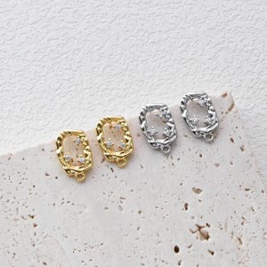 May include: Two pairs of earring findings, one pair is gold and the other is silver. Both pairs have a rectangular shape with a textured edge and a small clear crystal in each corner.