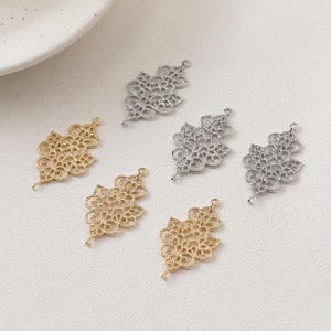 May include: A collection of six metal filigree charms in gold and silver tones. Each charm features an intricate, symmetrical, lace-like pattern with a small loop at the top and bottom for attachment, arranged in a scattered layout.