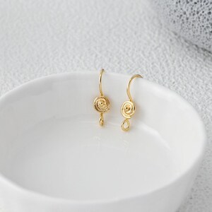 Può includere: Un paio di orecchini color oro con un design a spirale. Ogni orecchino presenta un gancio curvo e una piccola forma a goccia pendente. Gli orecchini sono esposti in una ciotola bianca.