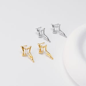 May include: Four earrings, two silver and two gold, are displayed on a white surface. Each earring features a rectangular, clear stone set within a starburst design. The silver earrings are on the top, and the gold earrings are on the bottom.