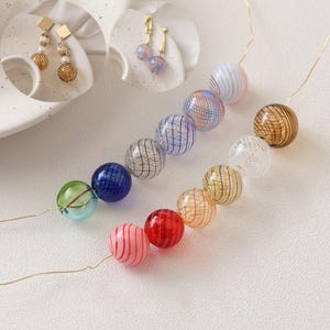 Könnte beinhalten: Eine Sammlung farbenfroher Glasperlen in verschiedenen Größen und Designs. Die Perlen sind auf dünnem Golddraht aufgefädelt und weisen gestreifte und gemusterte Details in Blau-, Rot-, Gelb- und Grüntönen auf. Das Bild zeigt auch goldene Ohrringe.