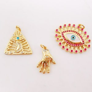 May include: Three gold-tone charms with an evil eye design. The first charm is a triangle with a crescent moon and an evil eye in the center. The second charm is a hand with an evil eye in the palm. The third charm is an evil eye with a white background and pink rhinestones around the eye.