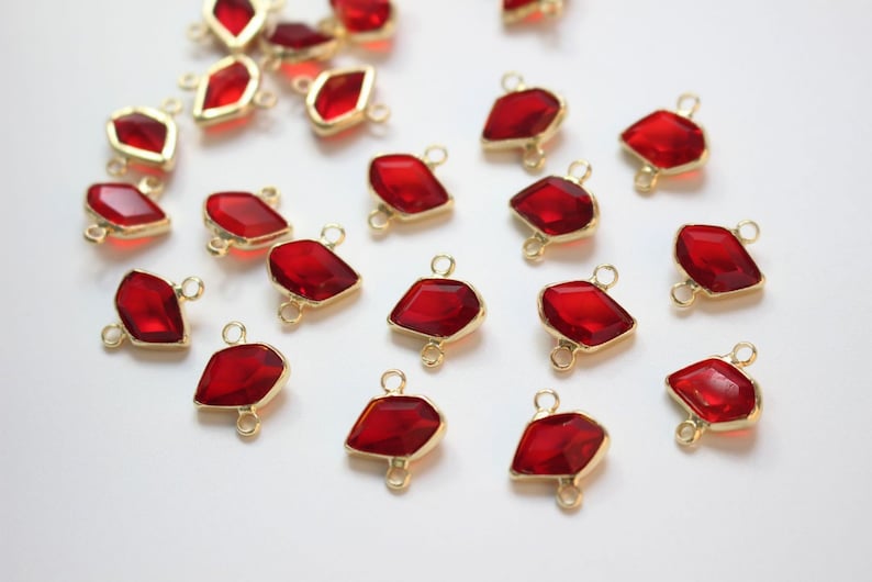 K&ouml;nnte beinhalten: Eine Sammlung von 20 roten facettierten Glas-Edelstein-Verbindern mit goldfarbenen Sprungringen. Die Verbinder sind tropfenf&ouml;rmig mit einer flachen R&uuml;ckseite und einer spitzen Spitze.