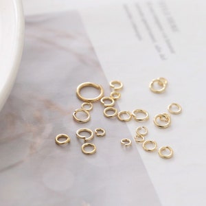 May include: A collection of gold-toned metal jump rings in various sizes, each featuring a small split opening. These circular jewelry findings are scattered across the surface, showcasing a polished metallic finish and a simple, open-ring design.