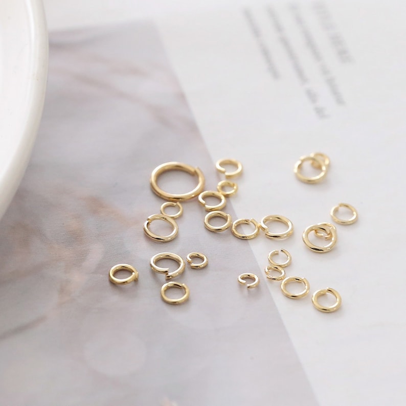 May include: A collection of gold-toned metal jump rings in various sizes, each featuring a small split opening. These circular jewelry findings are scattered across the surface, showcasing a polished metallic finish and a simple, open-ring design.