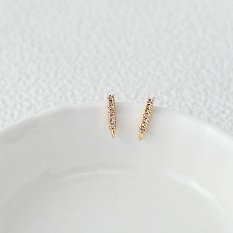 Puede incluir: Dos pendientes dorados en forma de barra, cada uno incrustado con peque&ntilde;as gemas transparentes. Los pendientes se exhiben sobre una superficie blanca, ligeramente curvada, creando una presentaci&oacute;n simple y elegante. Las gemas brillan.