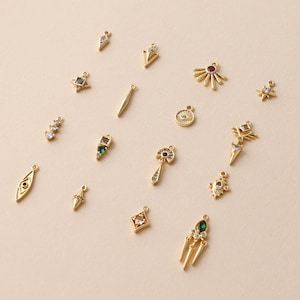 May include: A collection of small gold-toned metal charms featuring various geometric and symbolic shapes, including stars, arrows, eyes, and teardrops, accented with clear, green, and red embedded crystals and small jump rings for attachment.