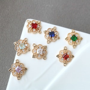 以下が含まれることがあります： 正方形のカラフルなラインストーンが中央にセットされた、ゴールドトーンの金属製チャームの詰め合わせ。チャームはラインストーンの周りに装飾的なフィリグリーデザインが施されています。チャームは白い表面の上に置かれています。