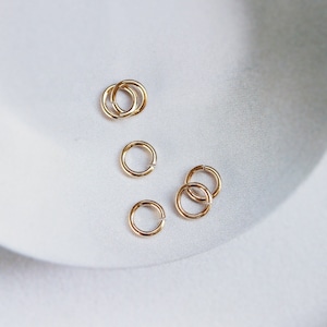 May include: A close-up of a group of small, gold jump rings. The rings are arranged in a loose cluster on a white surface.