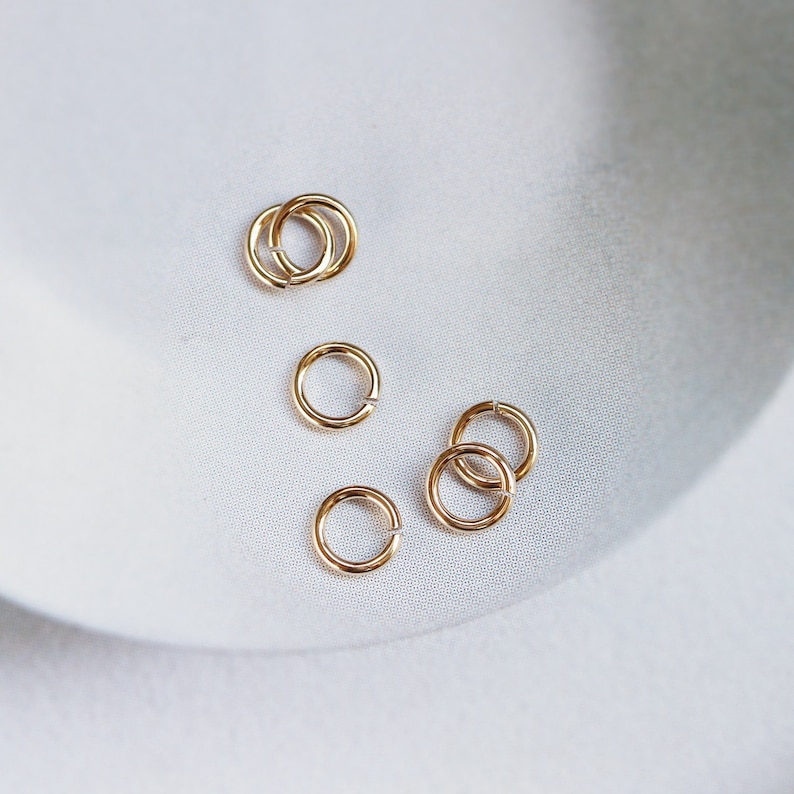 May include: A close-up of a group of small, gold jump rings. The rings are arranged in a loose cluster on a white surface.