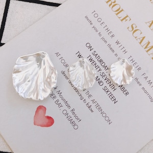 Könnte beinhalten: Zwei silberne Muschel-Anhänger aus Metall mit einem Loch zum Aufhängen. Die Anhänger befinden sich auf einem weißen Hintergrund mit dem Text "Gemeinsam mit ihrer Familie laden sie Sie ein, sich ihnen anzuschließen, um ihre Hochzeit zu feiern. Am Samstag, dem siebenundzwanzigsten und sechzehnten September, um vier Uhr nachmittags, mit anschließendem Abendessen und Tanz. K Mountain Resort, Cedar Bay, Ontario."