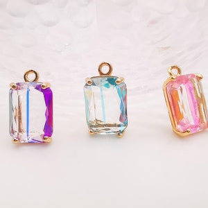 Op de afbeelding: Drie gouden hangers met heldere, gefacetteerde glazen stenen. De stenen zijn iriserend en hebben een regenboog effect. De hangers hebben de vorm van een rechthoek met afgeronde hoeken.