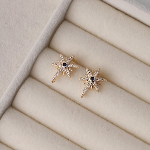 May include: A pair of gold-toned starburst earrings featuring a central dark blue stone surrounded by small clear pavé crystals. The star-shaped design has multiple radiating points of varying lengths, creating a symmetrical celestial appearance.