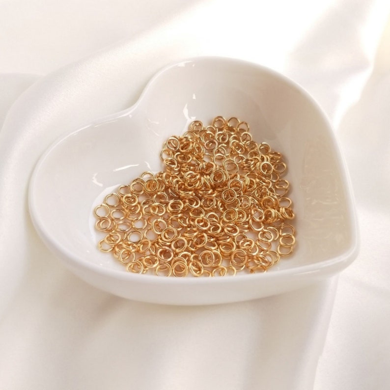 May include: A collection of small, circular gold-toned metal jump rings piled inside a white, heart-shaped ceramic dish. The rings are uniform in size and feature a simple, open-loop design, creating a dense cluster of metallic gold components.