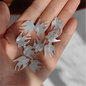 Könnte beinhalten: Eine Sammlung kleiner, schillernder weißer Perlmutt-Anhänger in Form von Schwalben. Jedes Stück weist ein detailliertes Ausschnittdesign mit gravierten Flügel- und Schwanzfedern sowie ein kleines Loch zur Befestigung auf und hat eine glatte Oberfläche.