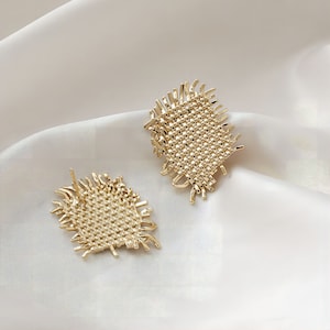Peut inclure: Une paire de boucles d'oreilles dorées avec un design tissé et texturé. Les boucles d'oreilles ont une forme géométrique carrée avec des bords effilochés.