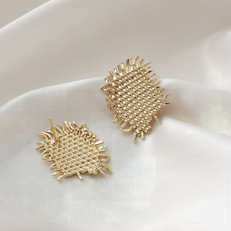 May include: A pair of gold-toned earrings with a woven, textured design. The earrings have a geometric, square shape with frayed edges.
