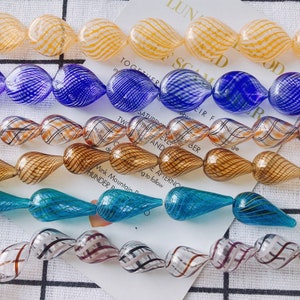 May include: Assortment of glass beads in various colours and patterns. The beads are teardrop shaped and have a swirl design. The colours include blue, brown, yellow, and turquoise.