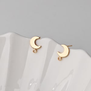 May include: A pair of gold-toned metal stud earrings, each shaped like a crescent moon. Each earring features a small, circular loop attached to the bottom of the moon, designed for hanging additional charms or decorative elements from the post.