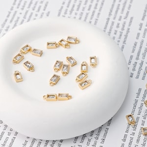 May include: Gold-colored rectangular charms with clear rectangular crystals are scattered on a white, rounded surface. Each charm has a small loop at each end. The charms are likely for jewelry making.