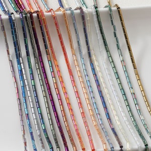 Könnte beinhalten: Sortiment an irisierenden Glasperlen in verschiedenen Farben, darunter Silber, Grün, Lila, Orange und Klar. Die Perlen sind klein und zylindrisch, mit einer glatten, glänzenden Oberfläche.