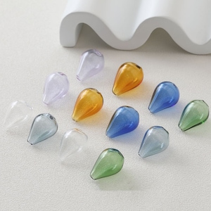 May include: A collection of teardrop-shaped glass beads in various colors, including clear, lavender, amber, blue, and green. The beads are arranged on a white surface, showcasing their translucent quality and potential for jewelry making or craft projects.