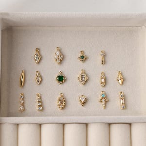 May include: A collection of fifteen small gold-toned metal jewelry charms arranged in three rows. The charms feature various geometric shapes, including diamonds, rectangles, and stars, accented with clear, green, and blue inset crystals.