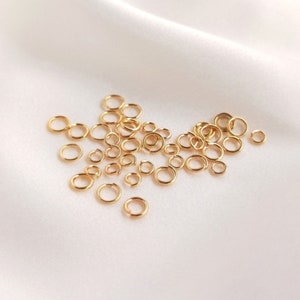 May include: A collection of small, circular gold-colored metal jump rings scattered across the frame. Each individual ring features a thin wire construction with a visible opening, designed for jewelry making and connecting various components.