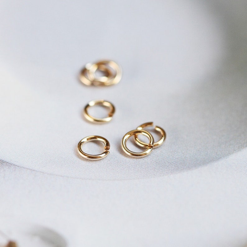 May include: A close-up of several small, gold-colored jump rings. The rings are arranged in a scattered pattern on a white surface.