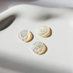 May include: Three iridescent white, square-shaped buttons with a raised unicorn design.