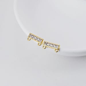 May include: Two gold-coloured bar-shaped jewellery pieces with clear rectangular stones. Each piece has two small loops on the ends. The jewellery is displayed on a white surface.