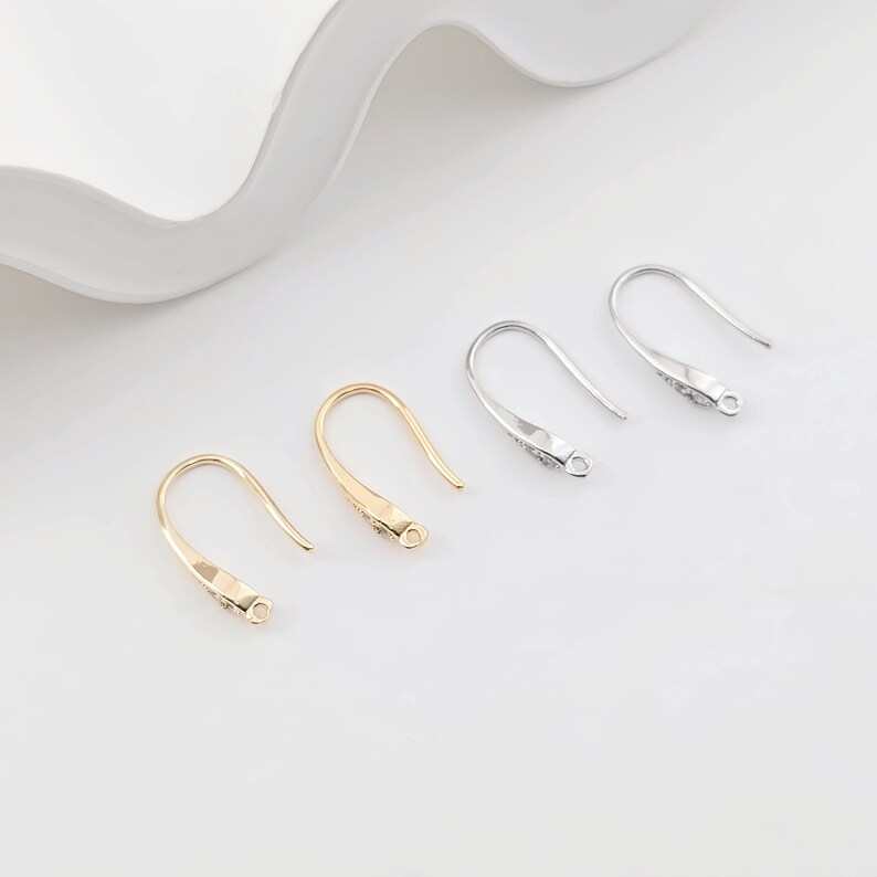 May include: Four earring hooks, two gold and two silver, are arranged in a row on a white surface. The hooks are all the same size and shape, with a small loop at the top and a curved hook at the bottom.
