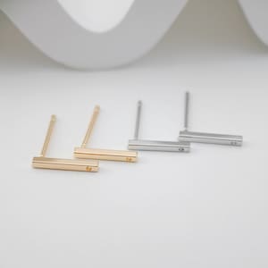 May include: Three pairs of minimalist bar earrings in gold, silver, and rose gold. Each earring features a thin, rectangular bar with a small, round stud in the center.