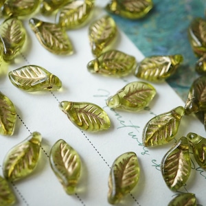 Può includere: Primo piano di perline di vetro a forma di foglia verde oliva con venature color oro. Le perline sono sparse su una superficie bianca con deboli linee tratteggiate e scritte a mano. Ideali per la creazione di gioielli e progetti artigianali.