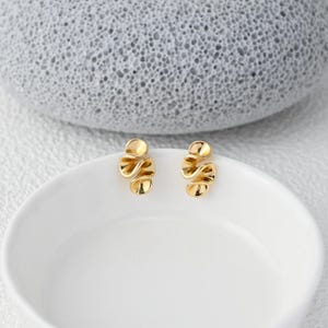 以下が含まれることがあります： ゴールドカラーのスタッドピアス。各イヤリングは、流れるようなレイヤー状の外観を持つ、ユニークな抽象的なデザインが特徴です。イヤリングは白い表面に表示され、背景には質感のある灰色の石があります。