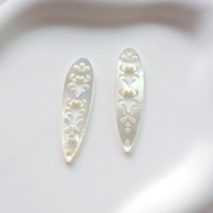 May include: Two white mother-of-pearl shell pendants with a carved floral design. The pendants are oval shaped and have a smooth, polished finish.