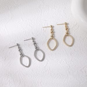 May include: Two pairs of earrings, one silver and one gold, are displayed on a white surface. Each earring features a post, a chain of small beads, and an oval-shaped drop. The gold earrings have a textured oval, while the silver ones have a beaded oval.