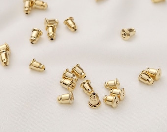 May include: A close-up of a group of gold-colored earring backs. The backs are small and round, with a small hole in the center. They are scattered on a white surface.