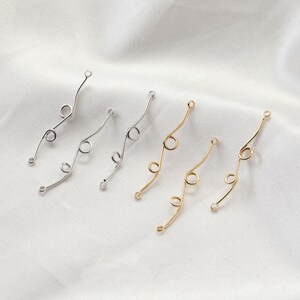 May include: Four silver and gold metal jewellery findings with a curved design and multiple loops. The findings are arranged in a row on a white surface.