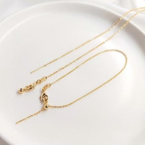 Peut inclure: Un collier en cha&icirc;ne dor&eacute;e avec une petite boule &agrave; l'extr&eacute;mit&eacute; de la cha&icirc;ne. La cha&icirc;ne est sur une surface blanche.