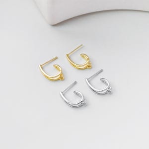 May include: Four pairs of earrings in gold and silver tones are displayed on a white surface. The earrings have a curved design with a post for piercing and small loops at the bottom. The gold earrings have a shiny finish, while the silver ones have a matte appearance.